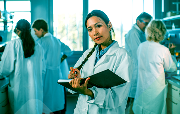 A researcher holding an open notebook and a pen looks up from her notes and out of the screen. The lab is busy with four other researchers behind her.